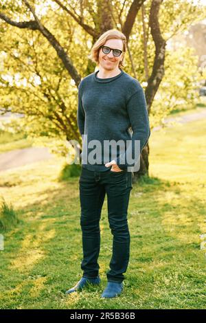 Ritratto all'aperto di bell'uomo di 35 - 40 anni con capelli rossi, in posa in verde parco soleggiato, con indosso un pullover blu e occhiali da sole Foto Stock