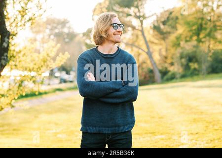 Ritratto all'aperto di bell'uomo di 35 - 40 anni con capelli rossi, in posa in verde parco soleggiato, con indosso un pullover blu e occhiali da sole, croci di armi Foto Stock