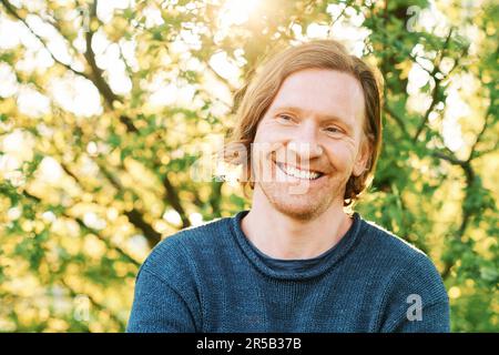 Ritratto all'aperto di bell'uomo di 35 - 40 anni con capelli rossi, in posa in verde parco soleggiato, indossando un pullover blu Foto Stock