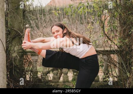 Felice insegnante di yoga femminile sorridente guardando la macchina fotografica in posizione estesa mano a piedi modificata. Indossando abiti casual sciolti bianchi e neri e calzino arancione Foto Stock