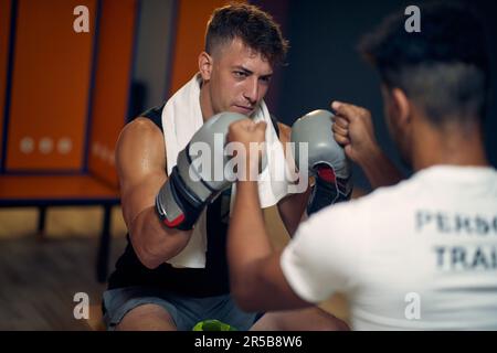 Deciso e focalizzato giovane pugilato sudato giocatore con il suo allenatore in spogliatoio seduto sulla panca, sensazione di potenza. Sport, stile di vita, arti marziali Foto Stock