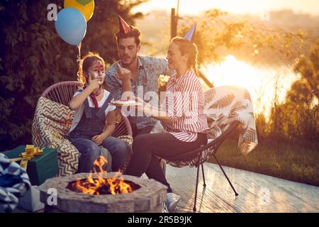 Felice famiglia che celebra il compleanno di figlia all'aperto in natura, seduta accanto al camino, torta di prova, compleanno ragazza con torta sul suo viso. Stile di vita, c Foto Stock