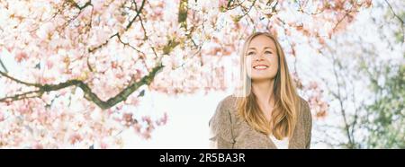 Sfondo banner panoramico con giovane bella donna felice con capelli biondi, posa sotto albero di magnolia fiorente Foto Stock