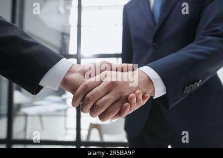 Uomini d'affari che scuotono le mani in piedi davanti alle finestre dell'ufficio Foto Stock