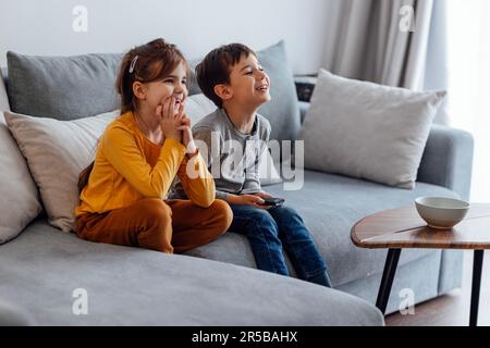 Due bambini sono seduti sul divano grigio e guardano la televisione. Ragazza carina e ragazzo ridendo e scegliendo cartoni animati in tv. Fratello e sorella spendono tim Foto Stock