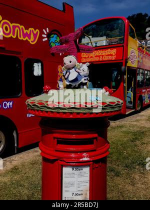 Lavorazione a maglia con scatola a lettere. Copertina in maglia a tema Alice nel paese delle meraviglie per la parte superiore della cassetta postale, autobus turistici a tema Alice per Conwy e Llandudno sullo sfondo. Foto Stock