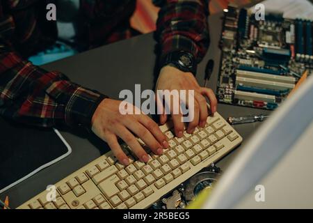 Visualizzazione ad angolo elevato della digitazione da parte di un giovane programmatore sulla tastiera del computer mentre si lavora con la scheda di sistema al tavolo Foto Stock