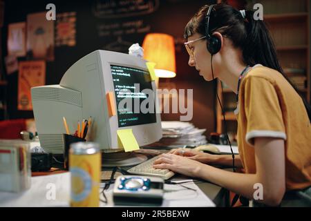 Giovane donna in cuffie digitando i codici per il nuovo software sul computer, che lavora nel suo posto di lavoro in garage Foto Stock