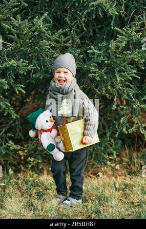 Ritratto all'aperto di felice dolce ragazzino in posa accanto al pino, con scatola regalo e giocattolo da snowman, con sciarpa di verme e cappello Foto Stock