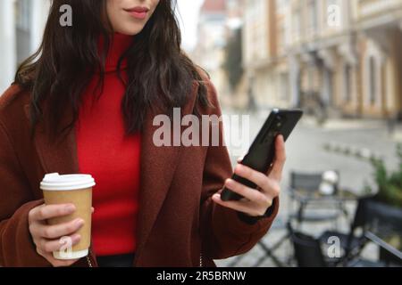 Donna usa il telefono cellulare all'esterno tenendo una tazza di caffè. ritagliare la foto Foto Stock