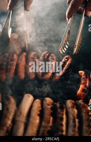 Un uomo con pinze in metallo sta rimuovendo con perizia i hot dog appena cucinati da una griglia a carbone Foto Stock