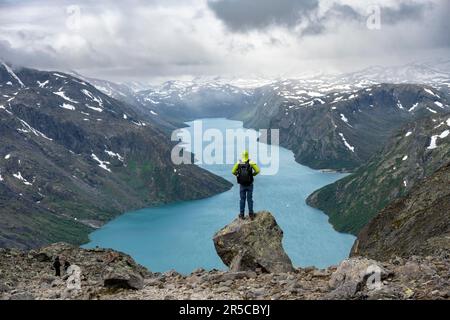 Arrampicatore in piedi sulle rocce, escursione Besseggen, passeggiata cresta, vista del lago Gjende e montagne innevate, Jotunheimen National Park, Vaga, Innlandet, Norvegia Foto Stock