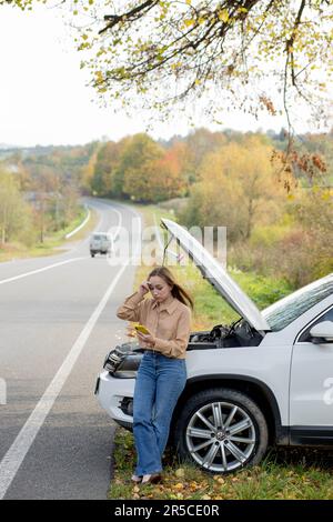 Una donna attende assistenza vicino alla sua auto rotta sul lato della strada. Donna con un'auto rotta con cofano aperto. Rovesciato giovane donna con telefono cellulare vicino auto rotta. Foto Stock