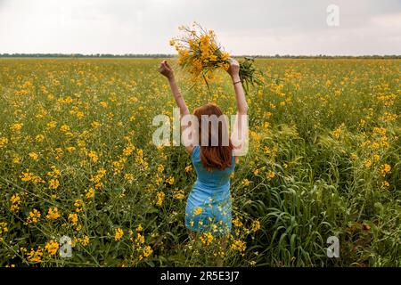 La ragazza tiene in mano un bouquet di fiori di colza gialli e spikelets verdi di grano sul campo Foto Stock