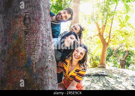 Un gruppo di amici diversi hanno un gioioso photoshoot nel parco. Nascondono giocosamente la testa dietro un grande albero, catturando momenti di lang candidi Foto Stock