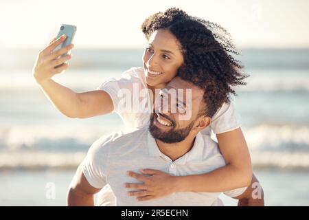 Coppia, piggyback e selfie in spiaggia, sorridere e felice in vacanza con sole estivo per blog, post e web. Uomo, donna e fotografia per romantici Foto Stock