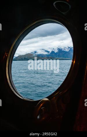 Vista panoramica attraverso il porthole di ottone del battello a vapore turistico che naviga sul Lago Wakatipu nell'Isola Sud della Nuova Zelanda Foto Stock