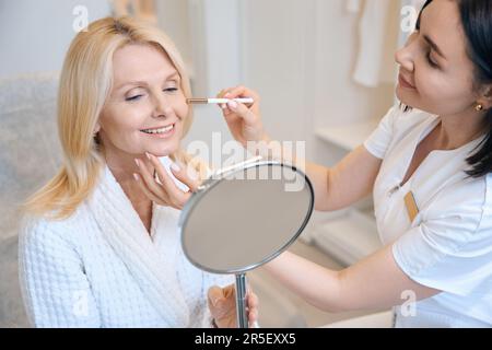 Il cosmetologo conduce le procedure per il cliente che si siede che osserva nello specchio Foto Stock
