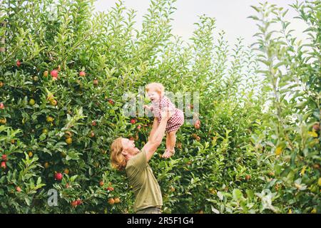 Felice giovane padre che gioca con adorabile bambina al di fuori, stile di vita familiare, papà tenendo il bambino in alto in aria, frutteto di mele Foto Stock