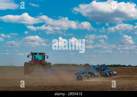Lavori agricoli nel campo. Il trattore rosso con l'aratro prepara il terreno sul campo agricolo con nuvole di polvere intorno Foto Stock