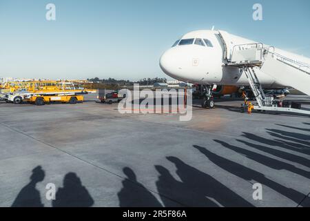 Nell'aeroporto di Lisbona, un aereo si trova sul lato destro con le scale. Le ombre delle persone formano una coda, assistite da una carrozza bagagli gialla. Lo sfondo è un bea Foto Stock