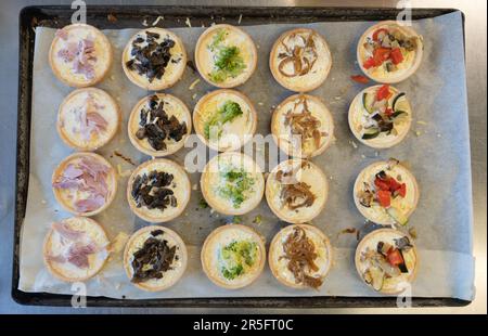 Deliziati con un mix di sapori con un assortimento di mini quiche, disposte in modo ordinato su un'ampia teglia da forno in metallo, pronta per la cottura alla perfezione. Foto Stock