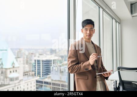 Giovane uomo d'affari asiatico che tiene un tablet guardando il pensiero della finestra. Foto Stock