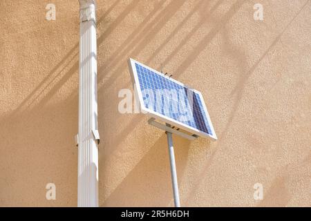 Un piccolo pannello solare montato su un palo metallico è posizionato accanto a una parete gialla dell'edificio, vicino a un tubo discendente dell'acqua piovana che si estende dal tetto Foto Stock