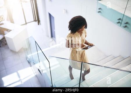 La tecnologia la aiuta a migliorare il suo gioco aziendale. una giovane donna d'affari che utilizza un tablet digitale mentre sale su una scala in un ufficio. Foto Stock