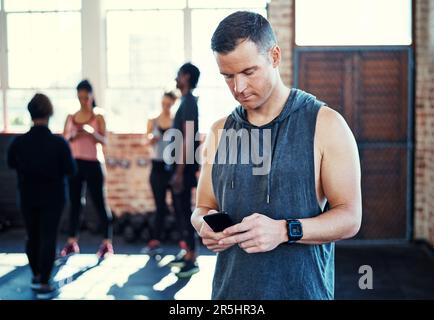 Ho avuto modo di tenere traccia dei miei progressi. un giovane uomo concentrato in piedi e utilizzando il suo cellulare prima di un allenamento in palestra. Foto Stock