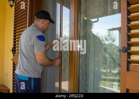 Immagine di un handyman che installa uno schermo del mosca in una finestra francese della sua casa. Protezione della casa da insetti. Foto Stock