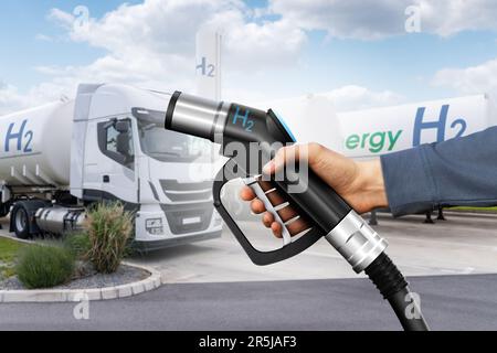 Mano con la pistola di rifornimento di idrogeno sullo sfondo dei rimorchi con serbatoio di idrogeno. Concetto di carburante pulito Foto Stock