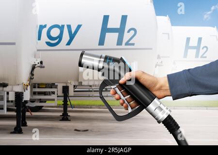 Mano con la pistola di rifornimento di idrogeno sullo sfondo dei rimorchi con serbatoio di idrogeno. Concetto di carburante pulito Foto Stock