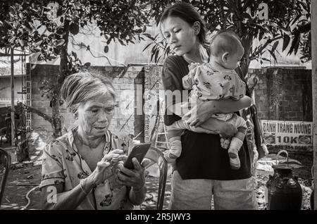 Una donna anziana della minoranza etnica del Brau usa il suo smartphone mentre è sorvegliata dalla sua nipote che tiene un bambino nel loro villaggio di Dak Me, Foto Stock