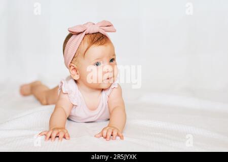 Il ritratto di adorabile bambino dai capelli rossi sdraiato sul ventre, su sfondo bianco, indossando il corpo rosa e la fascia, guarda a lato Foto Stock