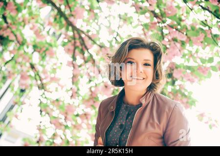 Ritratto primaverile all'aperto di una donna sorridente e felice che guarda giù per la fotocamera, fiori rosa in fiore sullo sfondo, indossando una giacca di pelle marrone Foto Stock