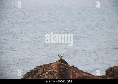 L'uomo avventuroso che si erge sul bordo di una scogliera gode della vista della costa atlantica nella regione di Odemira del Portogallo sudoccidentale. Vagando per la Ro Foto Stock