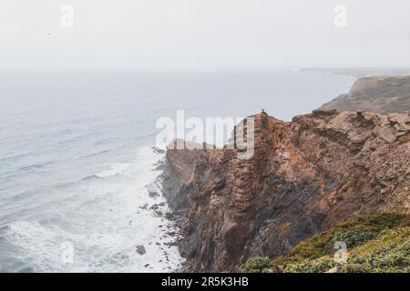 L'uomo avventuroso che si erge sul bordo di una scogliera gode della vista della costa atlantica nella regione di Odemira del Portogallo sudoccidentale. Vagando per la Ro Foto Stock