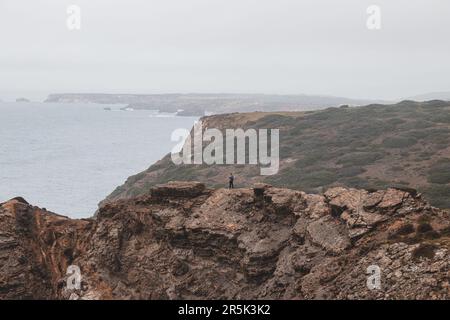 L'uomo avventuroso che si erge sul bordo di una scogliera gode della vista della costa atlantica nella regione di Odemira del Portogallo sudoccidentale. Vagando per la Ro Foto Stock