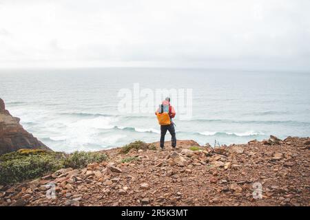 L'uomo avventuroso che si erge sul bordo di una scogliera gode della vista della costa atlantica nella regione di Odemira del Portogallo sudoccidentale. Vagando per la Ro Foto Stock