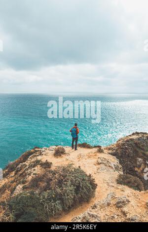 Avventuriero con uno zaino si erge sul bordo di una scogliera e osserva il bel gruppo di rocce giallo-oro di Punta de la Piedad a Lagos, nel Foto Stock