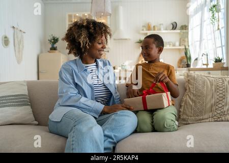 Sorridente entusiasta madre afro-americana che riceve un regalo di compleanno da un figlio a casa. Foto Stock