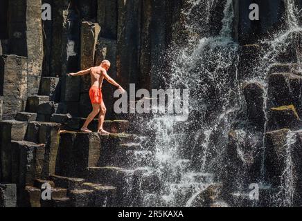 Uomo caucasico in costume da bagno in equilibrio vicino ai corsi d'acqua che cadono sulle cascate di pietra vulcanica nera. Cascata di Rochester Falls - popolare touri Foto Stock
