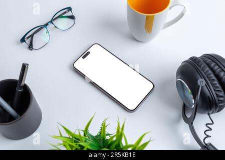 Mockup telefonico con schermo isolato per la presentazione delle applicazioni collocato sulla scrivania dell'ufficio, circondato da tazza da caffè, bicchieri, penne, piante e cuffie. Foto Stock