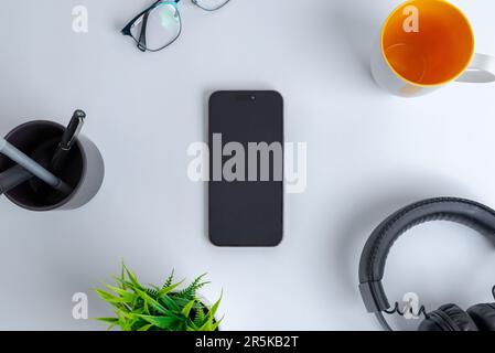 Telefono sulla scrivania. Vista dall'alto, composizione di giacitura piatta. Circondato da tazza di caffè, cuffie, piante, bicchieri e penne Foto Stock