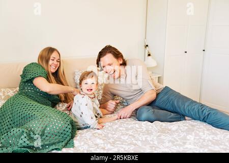 Giovane coppia felice madre e padre che gioca con adorabile bambina di 1 anni, la famiglia si rilassa a letto Foto Stock