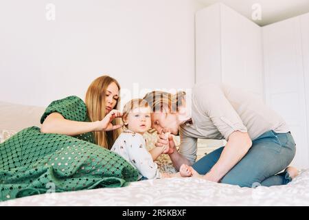 Giovane coppia felice madre e padre che gioca con adorabile bambina di 1 anni, la famiglia si rilassa a letto Foto Stock