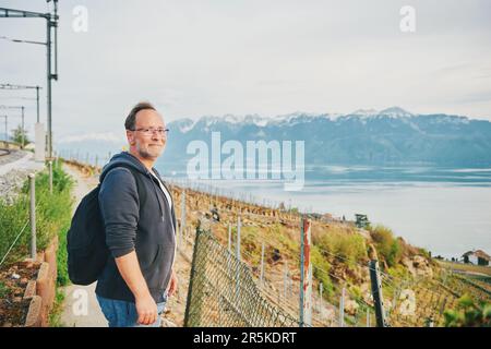 Uomo di mezza età trekking con zaino, in posa accanto alle ferrovie con il lago di Ginevra e Haute-Savoie vista, Lavaux, Svizzera Foto Stock