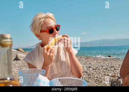 Picnic estivo sulla spiaggia. Gruppo di amici che hanno pic-nic con frutta fresca e antipasti all'aperto in riva al mare in spiaggia. Donna bionda che mangia la fetta di yel Foto Stock