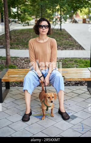 Giovane adulta in città con un cane seduto e abbracciato, sorridente. Accarezzare il cane, nutrirsi, divertirsi Foto di alta qualità Foto Stock